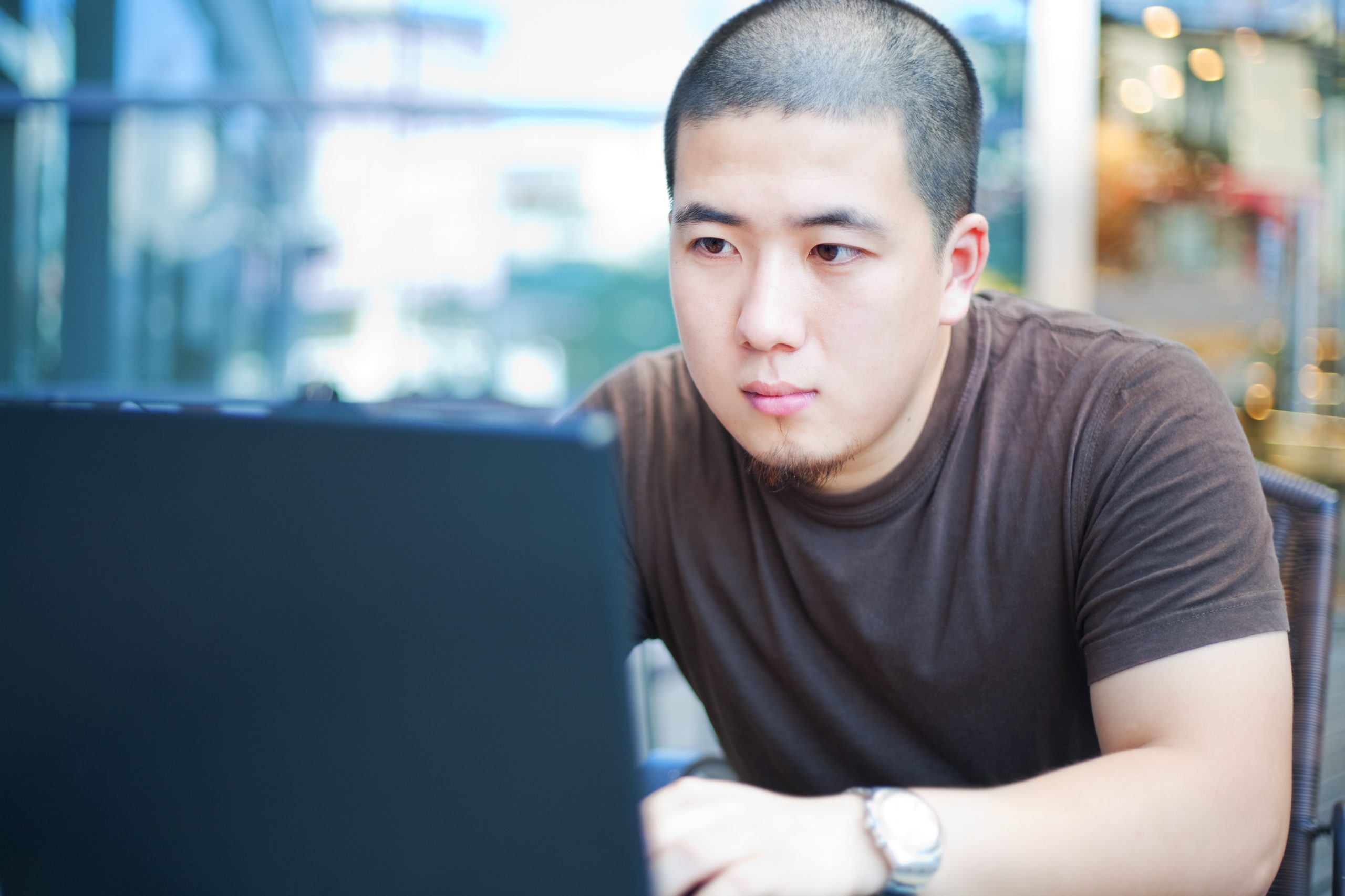 An asian young man is using a laptop in a outdoors cafe.